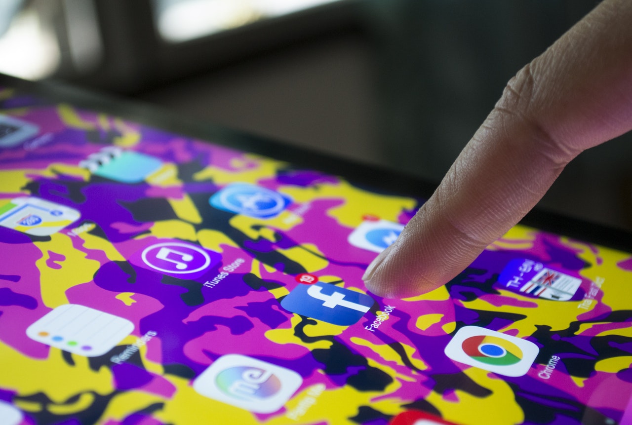 A mobile screen with a yellow, purple and pink background. A finger hovers over the Facebbok app while there is also a Chrome app and an iTunes app 