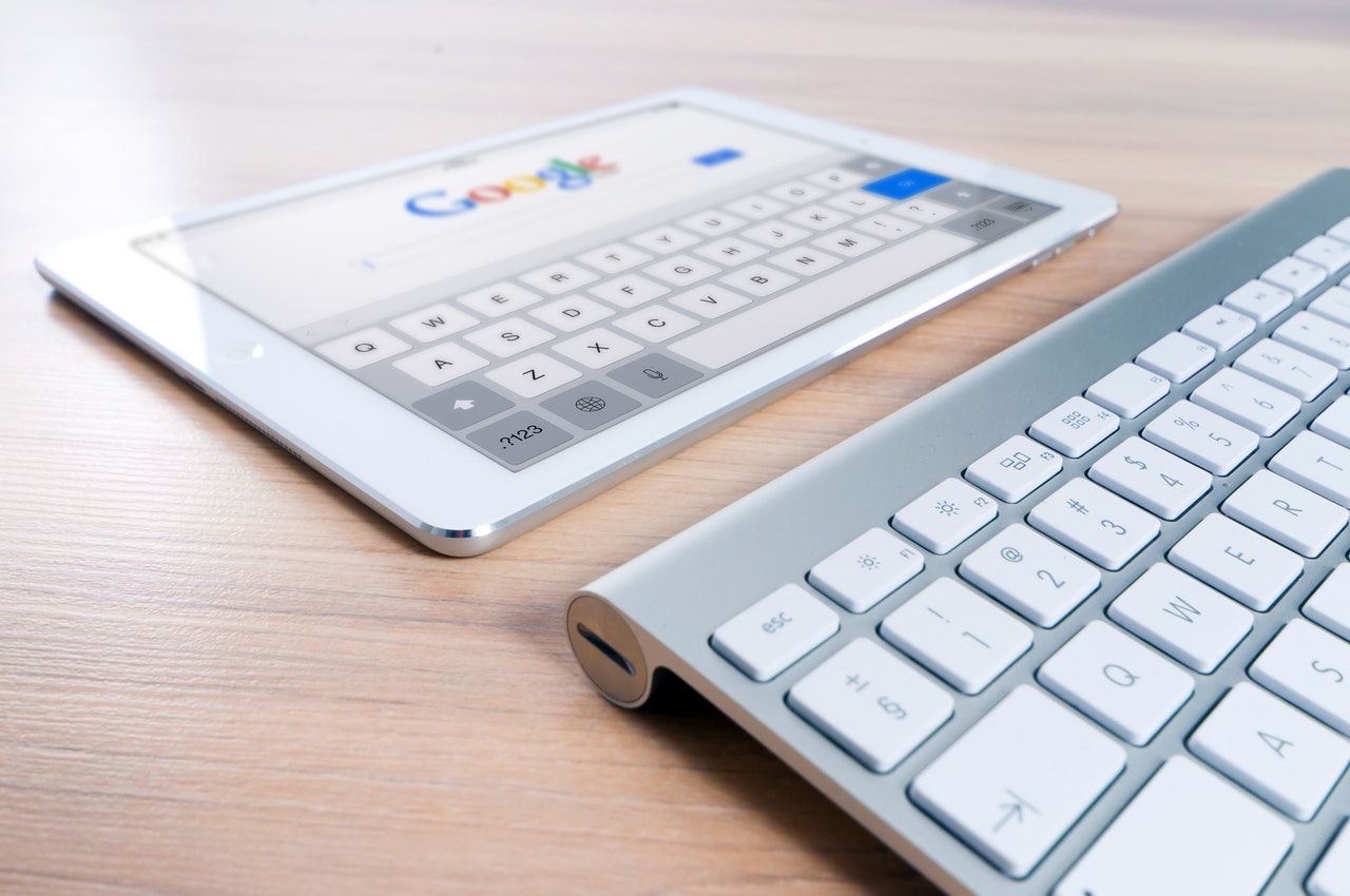 A tablet placed horizontally onto a wooden tablet. There is also a keyboard beneath the tablet, and the tablet is opened up on the Google search bar 