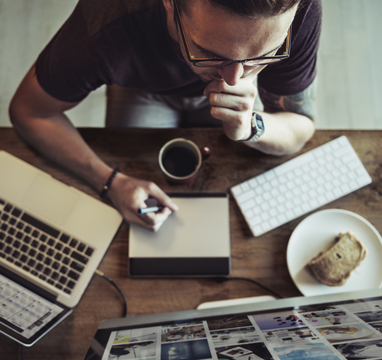 A birds-eye-view of a man sat at his desk looking at a big computer screen. He has a laptop beside him, a wireless keyboard, a cup of coffee and is using a digital pad and pen. 