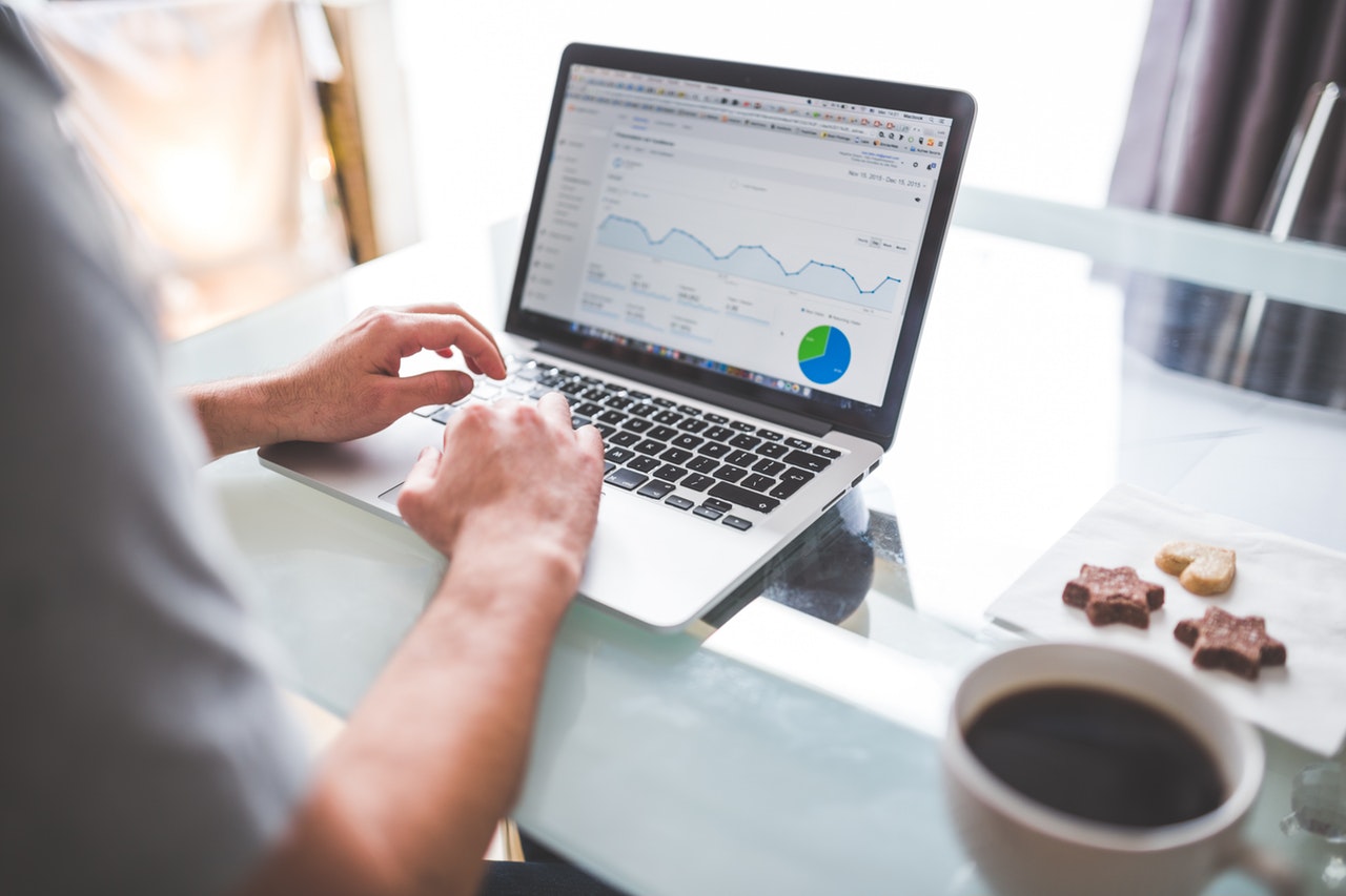 A man sitting on a laptop looking at a screen of statistics. There is a coffee beside him and some biscuits