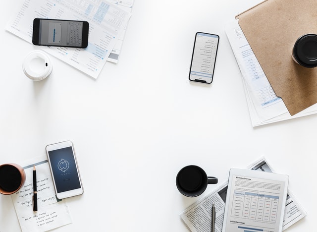 A bird's eye view of a table with mobile phones, notepads, coffee, pens and spreadsheets