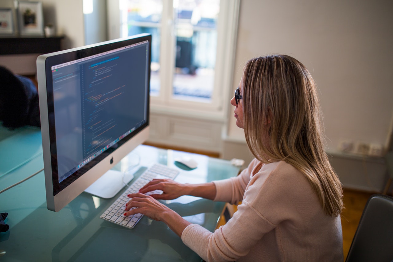 A woman sitting at a computer typing on her keyboard. She is wearing glasses and a cream jumper.