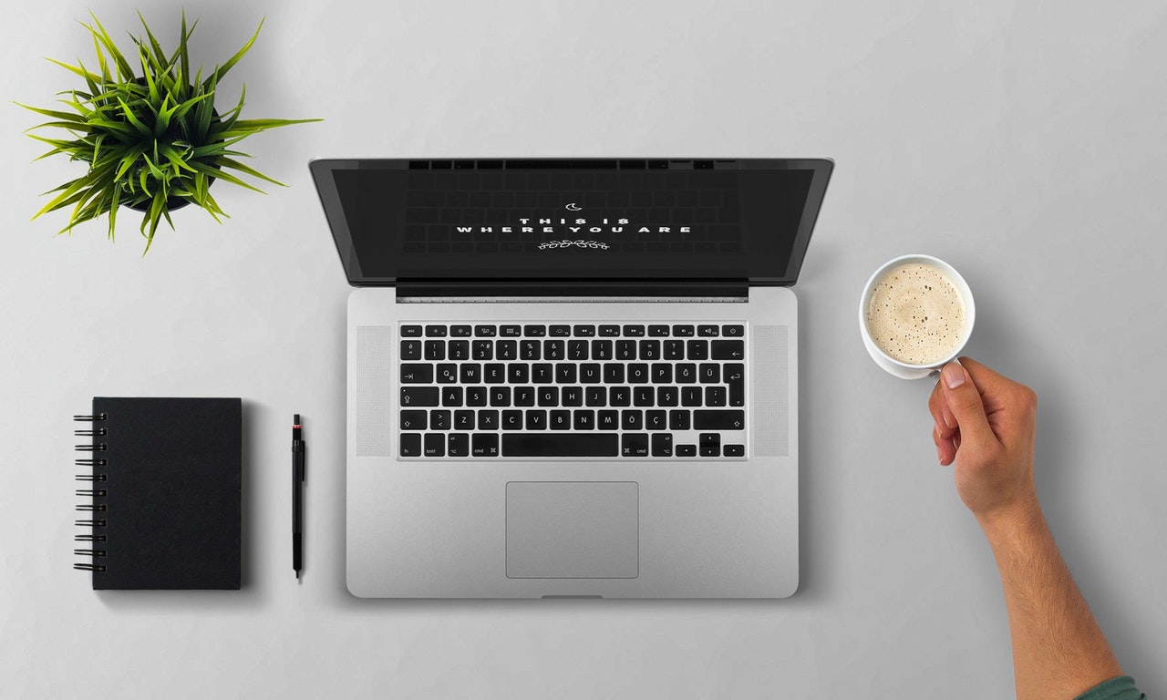 A birds-eye view of an open laptop with a plant and notebook to the left, and a person holding a coffee mug to the right 