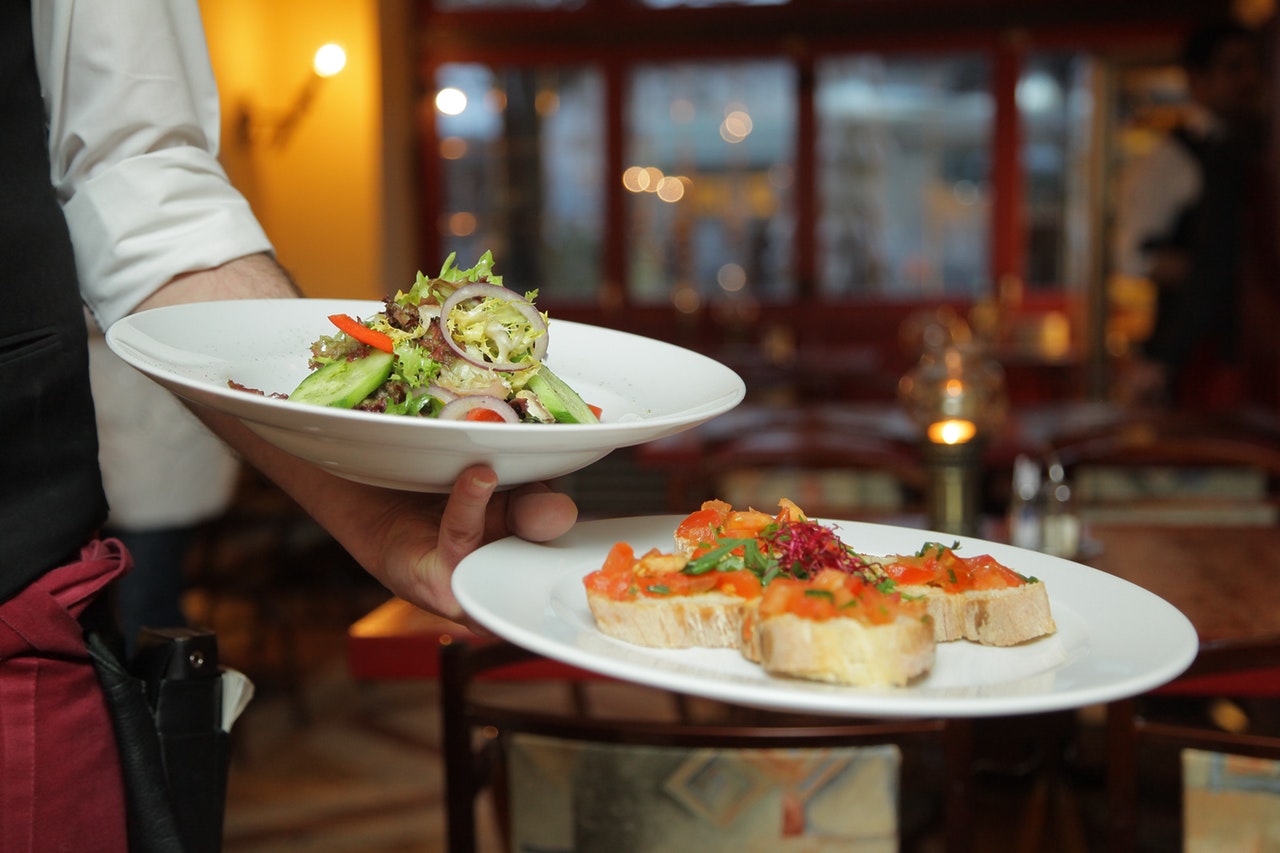 A waiter holding a plate of food in his left hand. He is also balancing a bowl of salad on his left arm 