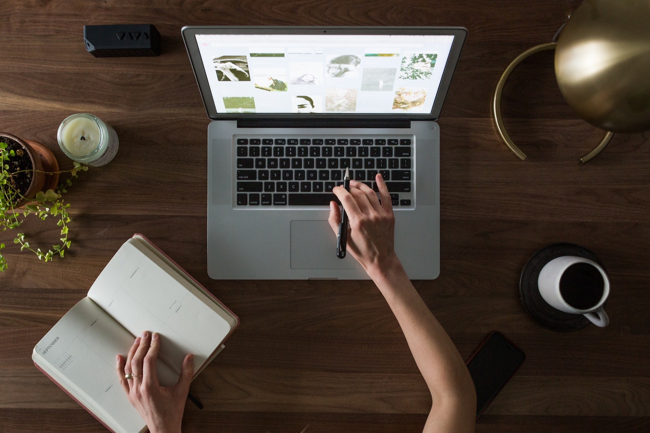 Bird's-eye-view of a lady sitting at a laptop with her left hand placed on a notebook and a coffee mug to her right. 