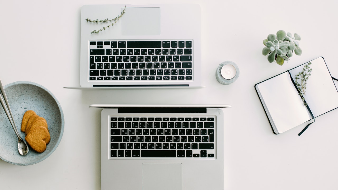 A birds-eye view picture of two laptops facing against one another. There is a bowl of heart-shaped cookies to the left and a notebook to the right of the image opened up to blank pages