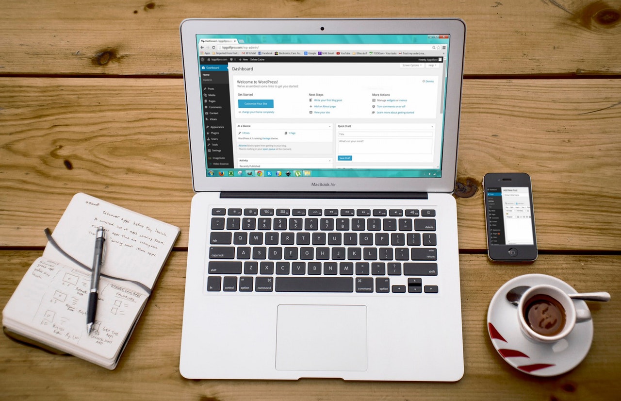 A laptop displaying the WordPress dashboard. A notebook and pen are to the left of the laptop. An espresso and mobile to the right. 