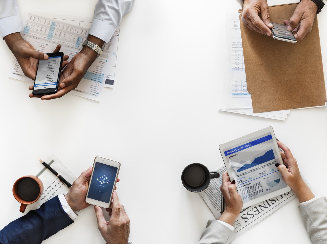 Bird's eye view of three people sitting at a table with mobile phones in their hands, and a fourth person holding a tablet Bird's eye view of three people sitting at a table with mobile phones in their hands, and a fourth person holding a tablet