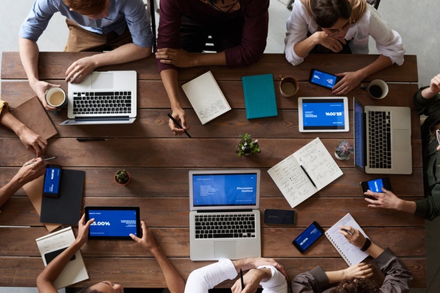 Bird's eye view of a wooden table with 8 people gathered round on laptops, mobiles and tablets.