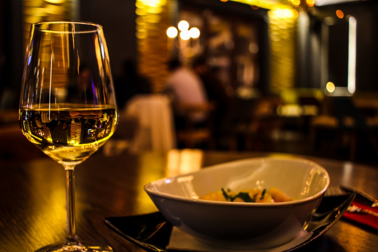 A close up of a wine glass and dish of food placed on a table. In the background is a restaurant.