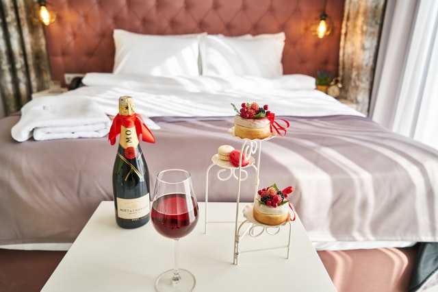 Image of a hotel bed with a small table featuring a bottle of champagne, glass of wine and some pastries